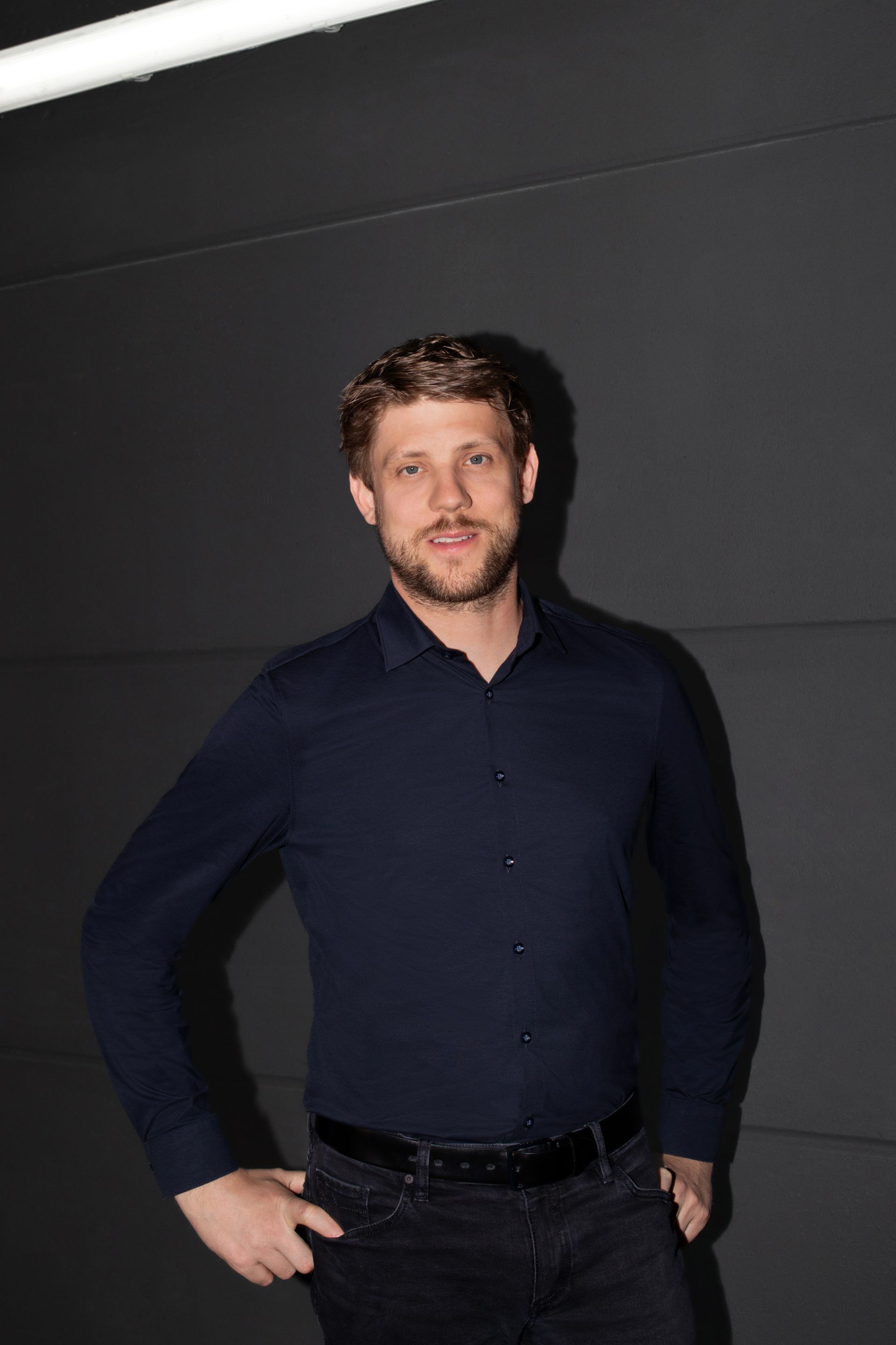 Muž oblečený v tmavo modrej košeli a čiernych nohaviciach stojí pred tmavým pozadím. Má zastrčené ruky do opaska. Pozadie je jednoduché a monochromatické.