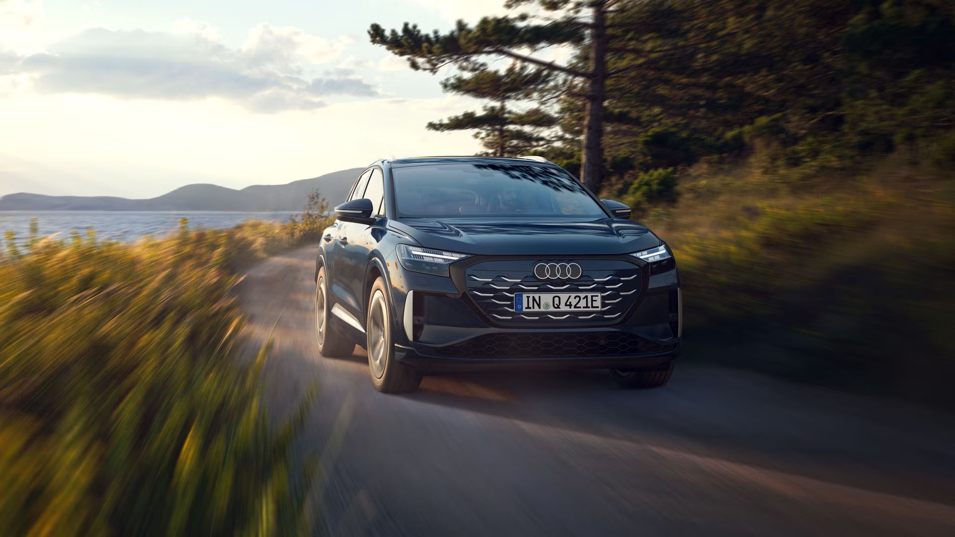 Q4 SUV e-tron z pohľadu na predné partie v plnej rýchlosti na ceste pri jazere, v pozadí je horská krajina.