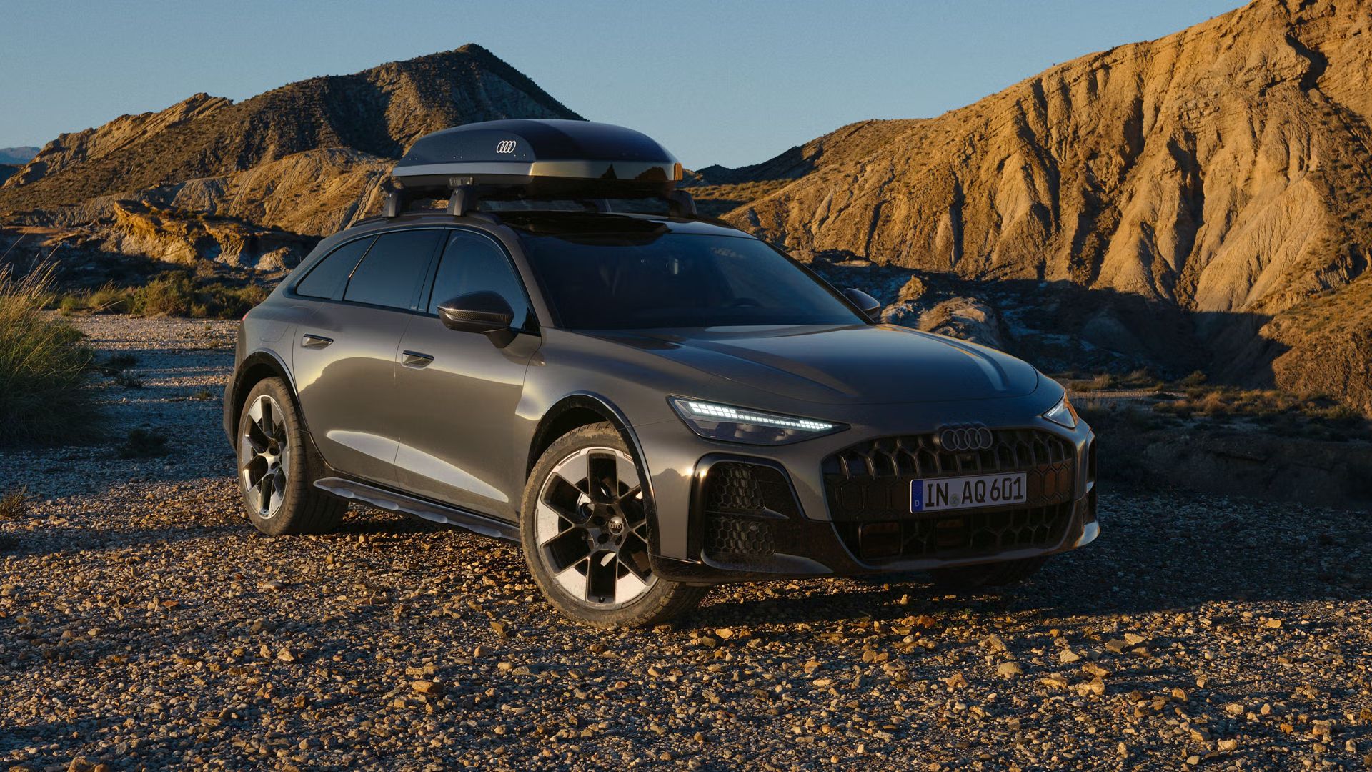 Audi A6 allroad, zachytené z čelného uhla pod uhlom, s čiernym strešným boxom v odtieni „brilliant black“, parkuje na štrkovej ceste. V pozadí sa rozprestiera pustá a skalnatá krajina, pričom bočné slnečné svetlo rozdeľuje scénu na tienisté a svetlé časti.