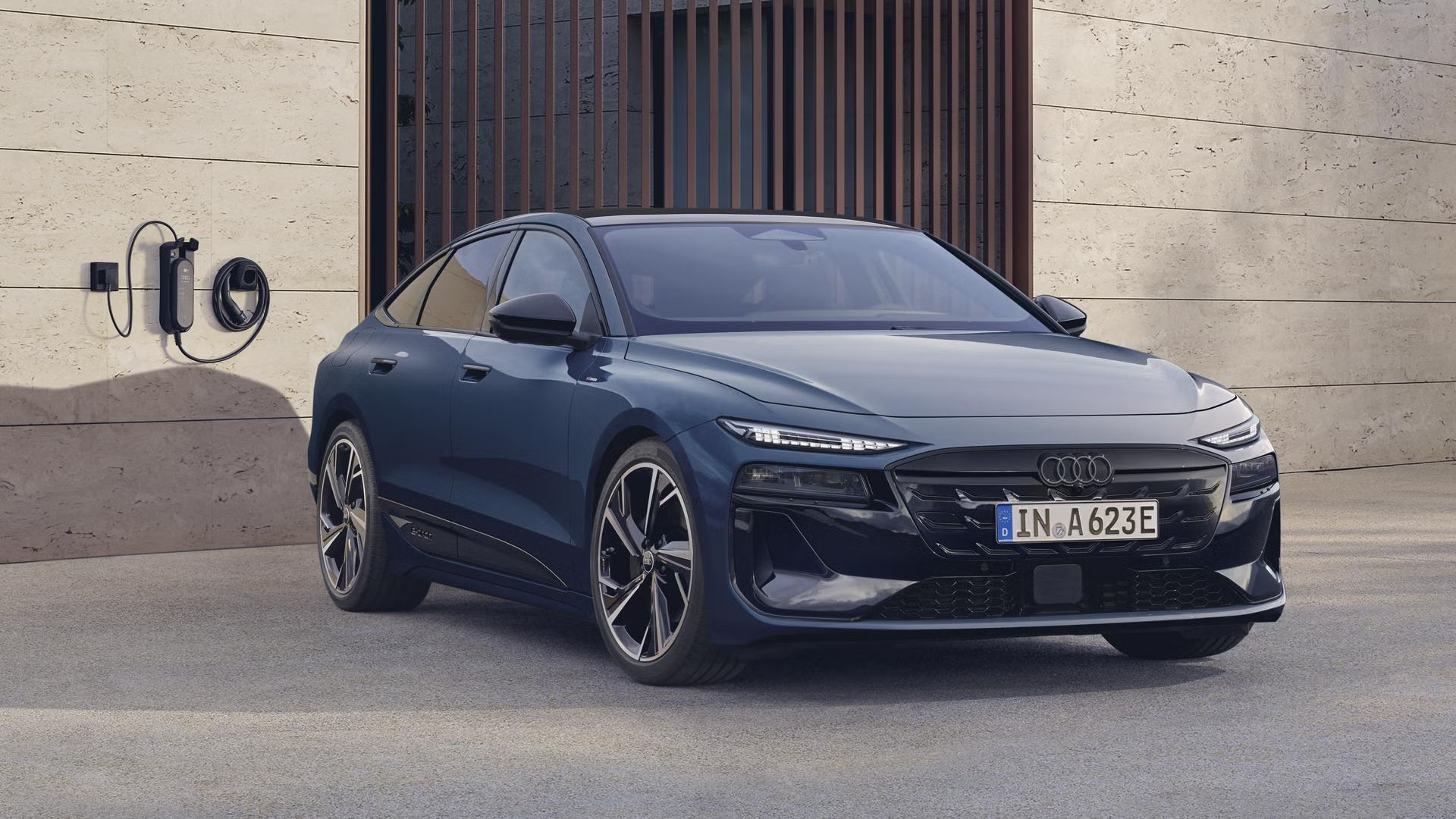 Šikmý predný profil modelu A6 Sportback e-tron, ktorý stojí na betónovom podklade pred budovou. Na fasáde budovy je namontovaná nabíjacie stanica. 