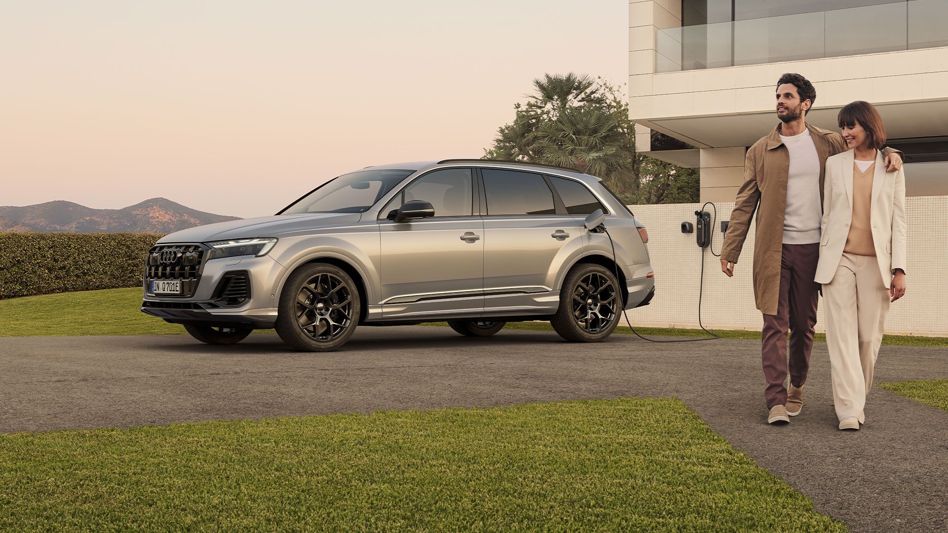 Bočný pohľad na strieborné Audi Q7 SUV TFSI e pri nabíjaní pri modernej vile. Okolo vozidla prechádza muž so ženou.