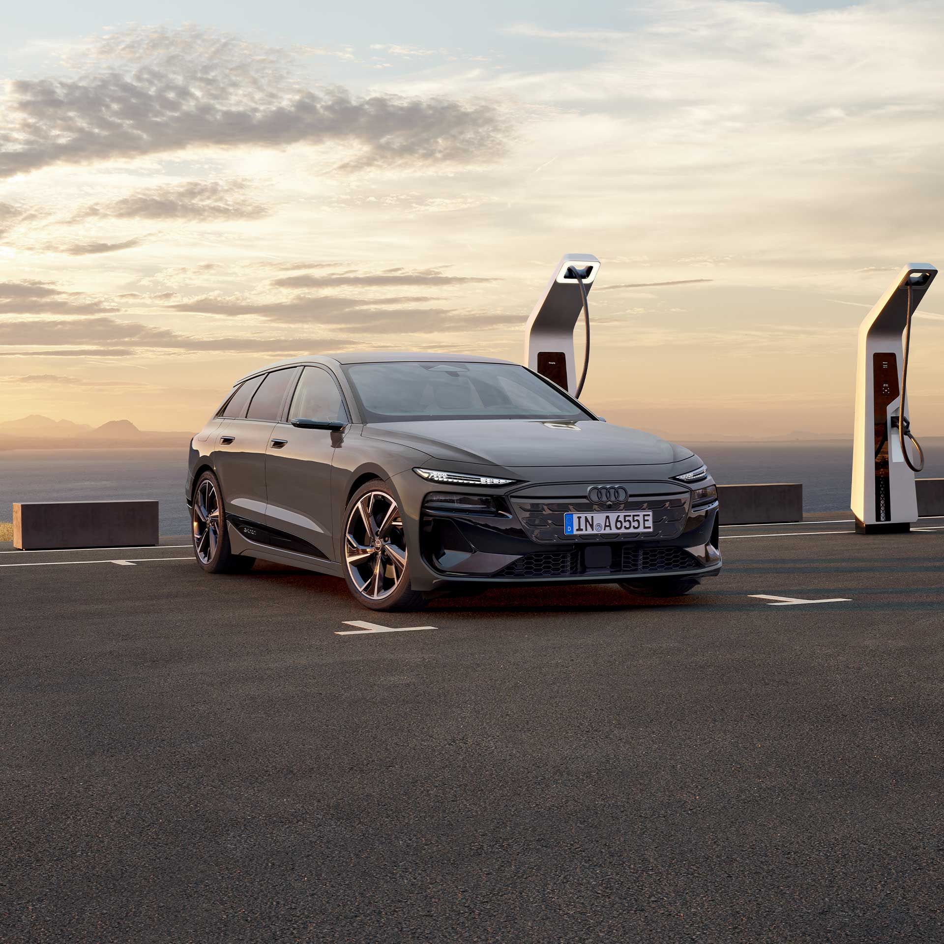 Audi A6 Avant e-tron zaparkovaný pri verejnej nabíjacej stanici na parkovisku pri pobreží. Za vozidlom je vidieť more a kopce v pozadí.