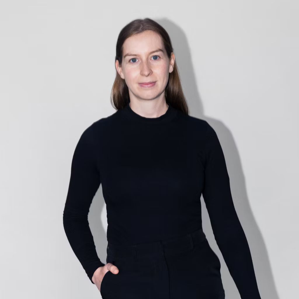 Žena v čiernom roláku pred bielou stenou, ktorá sa pozerá priamo do objektívu.