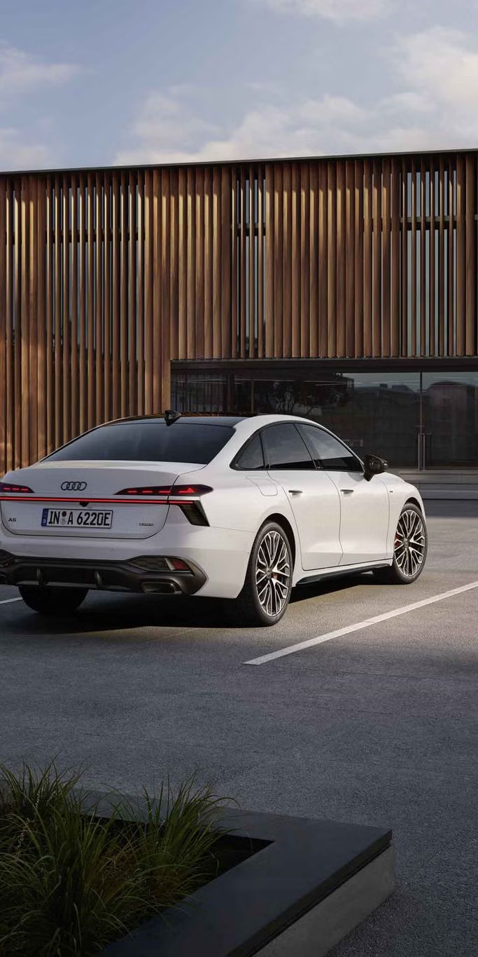 Die A6 Limousine e-hybrid in weiß steht auf einem Parkplatz vor einer Gebäudefassade. Eine junge Frau schließt die A6 Limousine e-hybrid an eine Ladestation an.