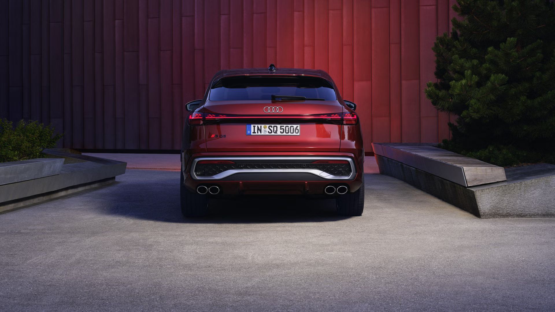 Pohľad na zadné partie Audi SQ5 Sportback zaparkované pred budovou s červenou fasádou. Vozidlo má zapnuté svetlá.