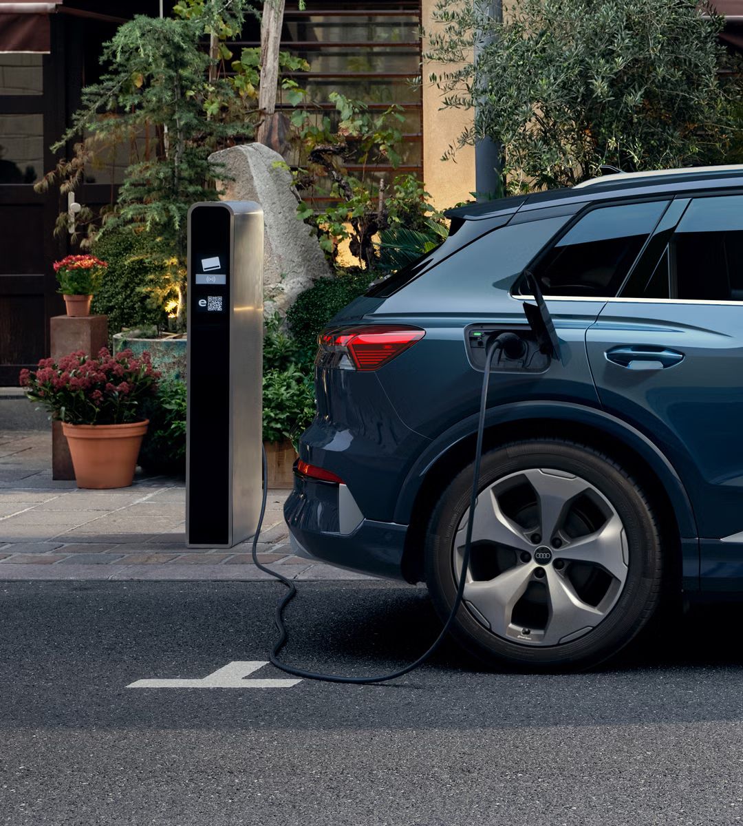 Detail na pravú zadnú časť Q4 SUV e-tron, ktoré parkuje pred budovou. Pred vchodom do budovy sú umiestnené kvety a stromčeky. Vozidlo je pripojené k verejnej nabíjacej stanici. 