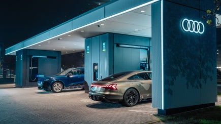 Audi charging hub v noci s osvetleným logo Audi kruhov a dvomi vozidlami zaparkovanými pri nabíjacích staniciach