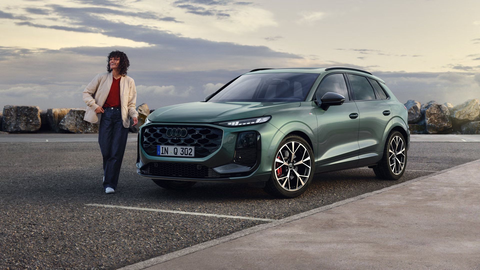 Bočný pohľad z prednej strany na zaparkované Q3 SUV na parkovisku pri pobreží, vedľa vozidla stojí mladá žena. 