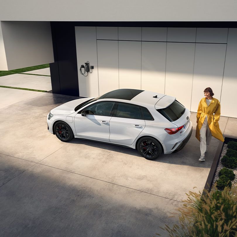 Audi A3 Sportback TFSI e zaparkované pre modernou budovou s nabíjacou stanicou na fasáde. Mladá žena prechádza okolo vozidla.
