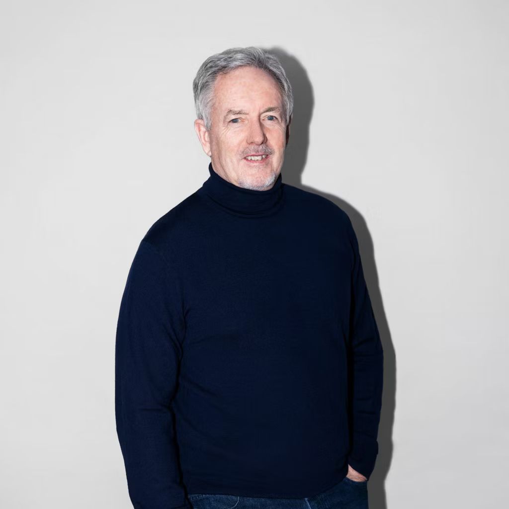 Muž v tmavomodrom roláku pred bielou stenou, ktorý sa pozerá priamo do kamery.