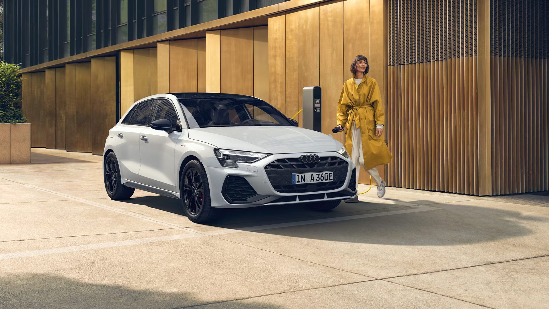 Audi A3 Sportback TFSI e zaparkované pri nabíjacej stanici pred modernou verejnou budovou. Pri vozidle stojí mladá žena a drží nabíjací konektor, ktorý chce zapojiť do vozidla.