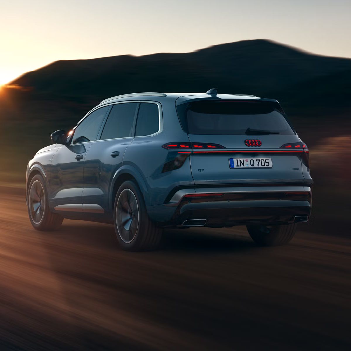 Audi Q7 SUV zachytený z boku zo zadnej strany pri plnej rýchlosti na vidieckej ceste, v pozadí sú hory. Scénu osvetľuje slnko zapadajúce za obzorom.  