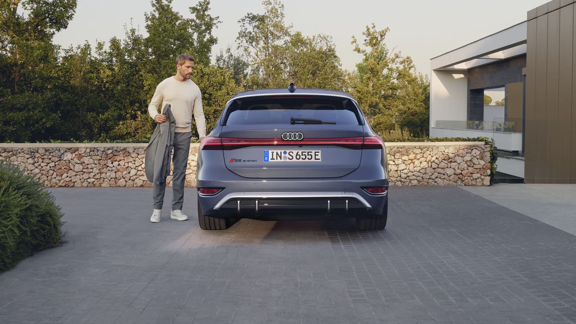Audi S6 Avant e-tron, ktoré stojí na dlažobných kockách pred kamennou stenou a záhradou moderného domu. Muž stredného veku sa chystá otvoriť dvere.
