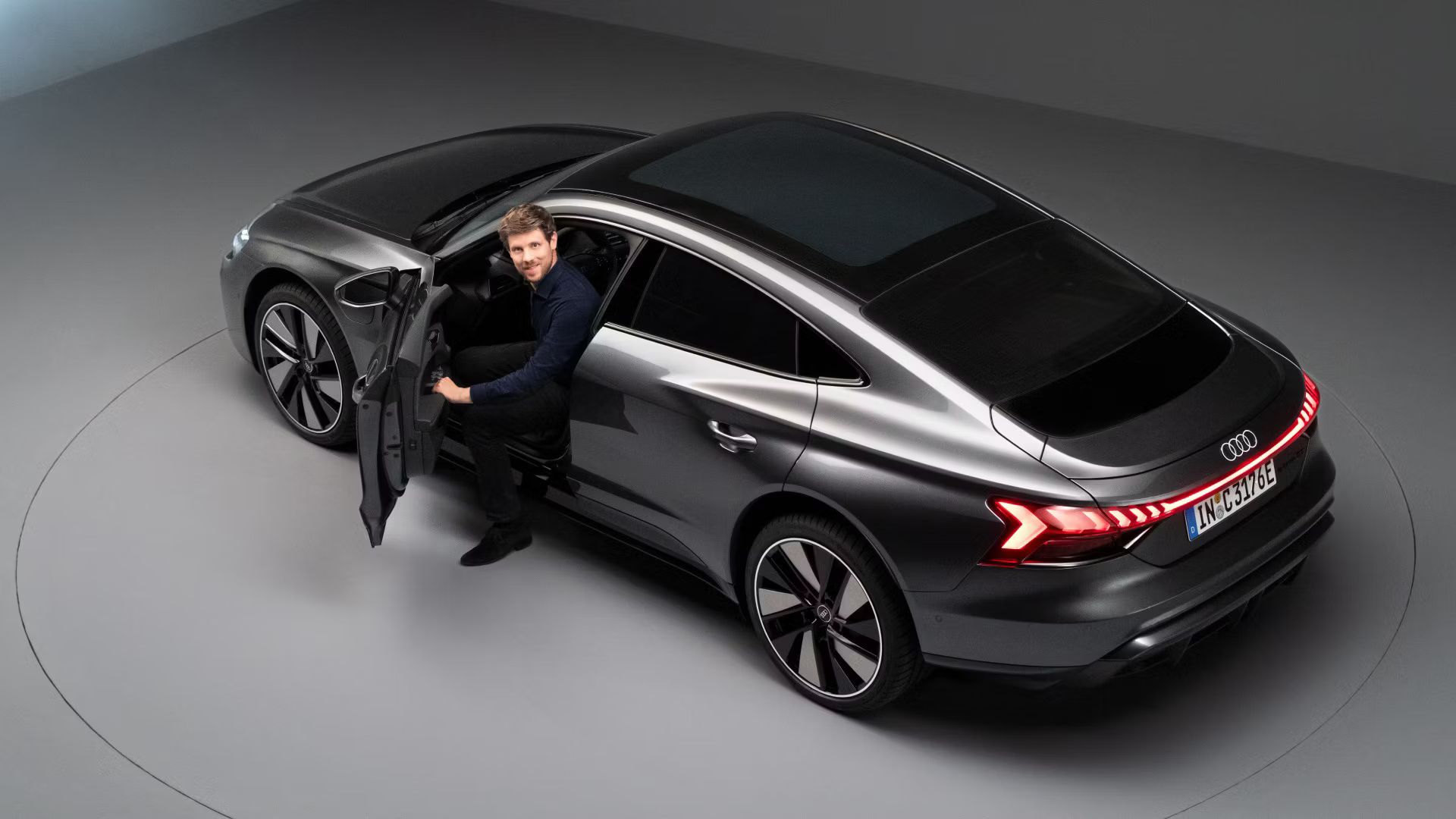 Čierne Audi e-tron GT quattro stojí na prezentačnom kruhu. Dvere vodiča sú otvorené, z vozidla vystupuje muž a pozerá sa priamo do kamery. Pohľad na zadné partie zhora.