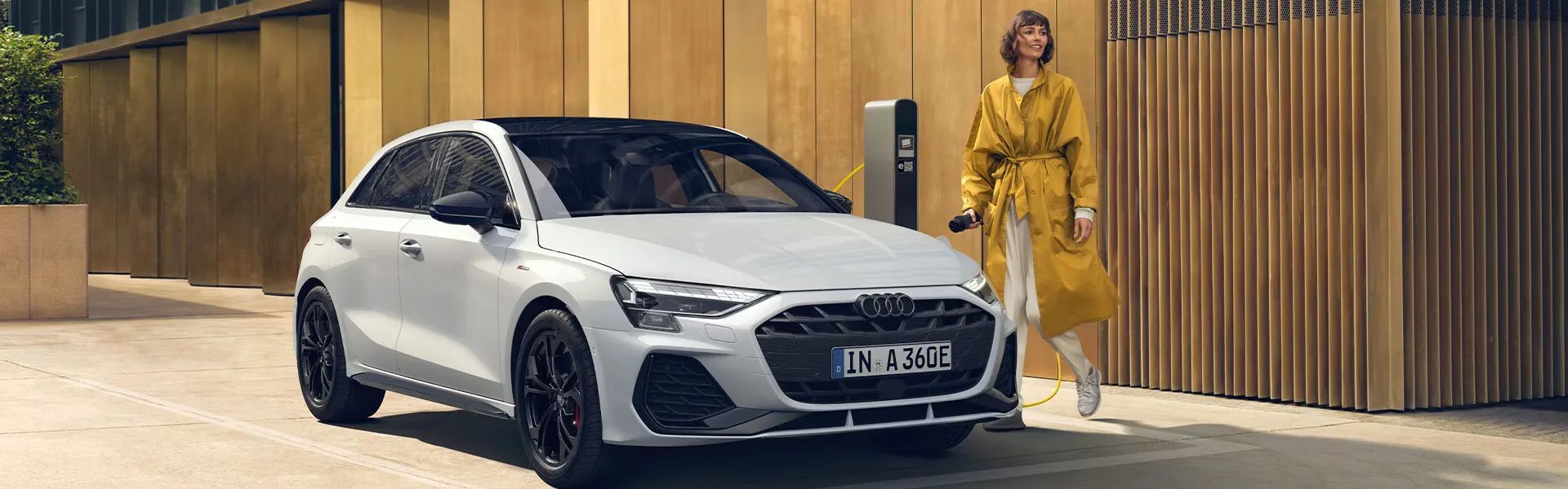 Audi A3 Sportback TFSI e je zaparkované pred modernou budovou pri nabíjacej stanici. Mladá žena v žltom jarnom kabáte drží v ruke nabíjací konektor a ide vozidlo pripojiť k nabíjacej stanici.