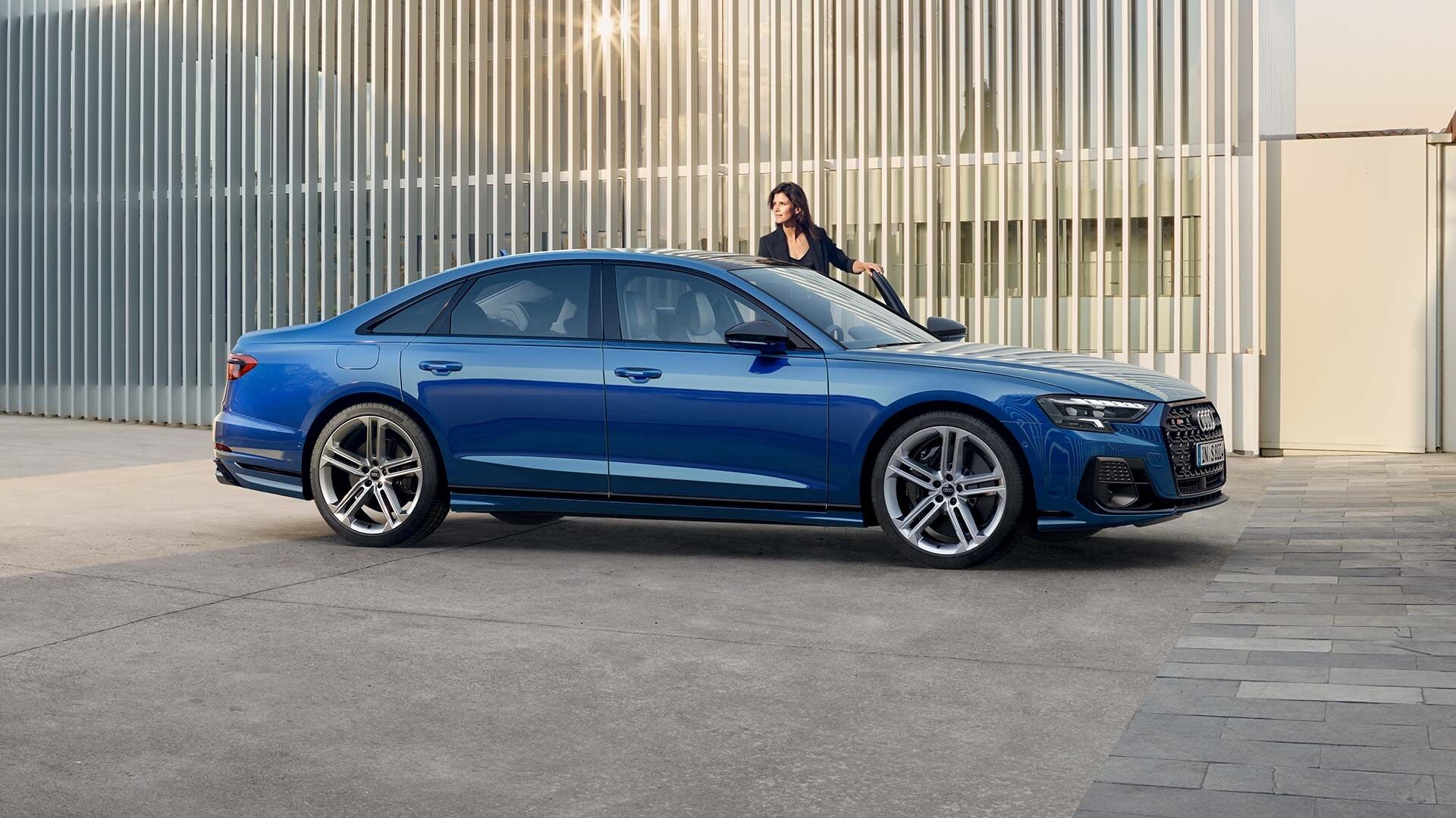 Modré luxusné Audi S8 je zaparkované pred modernou budovou. Pri aute stojí žena, ktorá sa opiera o predné dvere. V pozadí sú vertikálne biele lamely.