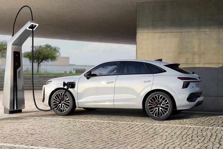 Audi Q3 Sportback e-hybrid zaparkované ori nabíjacej stanice vedľa modernej verejnej budovy. Vozidlo sa nabíja. V pozadí vidieť schody, stromy, zeleň a ďalšie budovy.