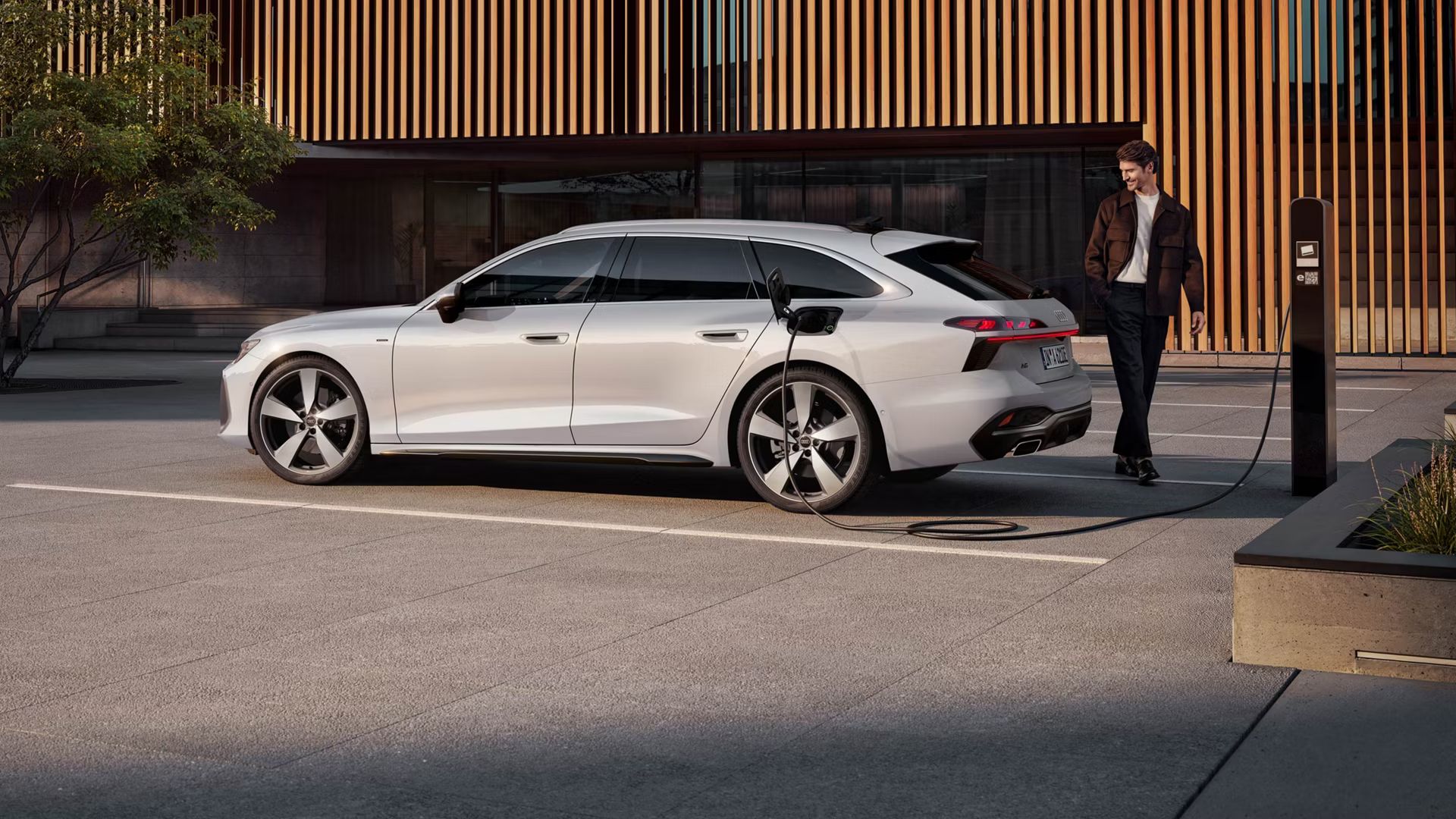 Audi A6 Avant e-hybrid je na parkovisku pripojený k nabíjacej stanici. Pohľad na vozidlo zozadu zboku.