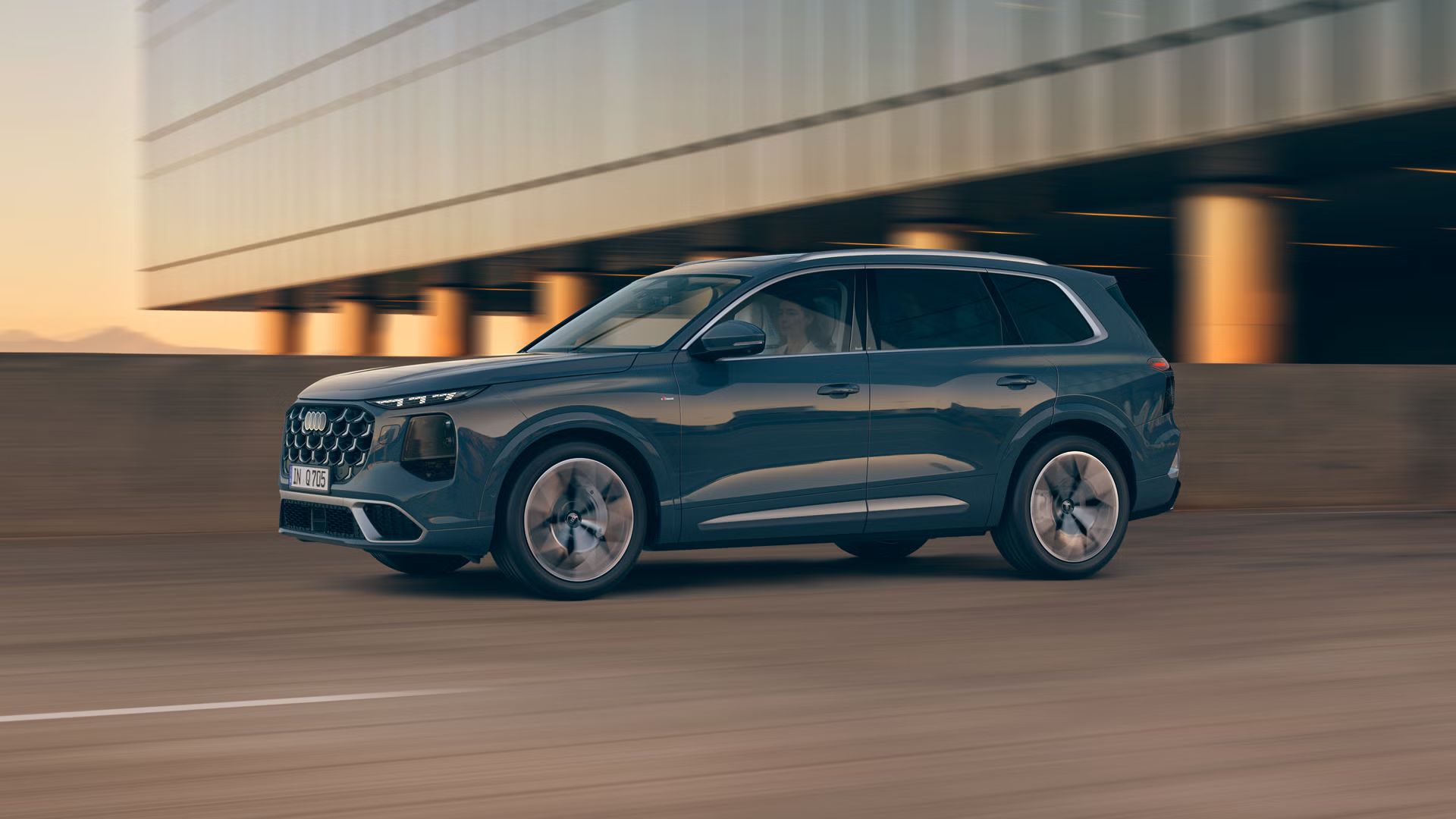 Modré SUV Audi Q7 ide po ceste pri západe slnka, v pozadí je moderná budova stojaca na betónových pilieroch.