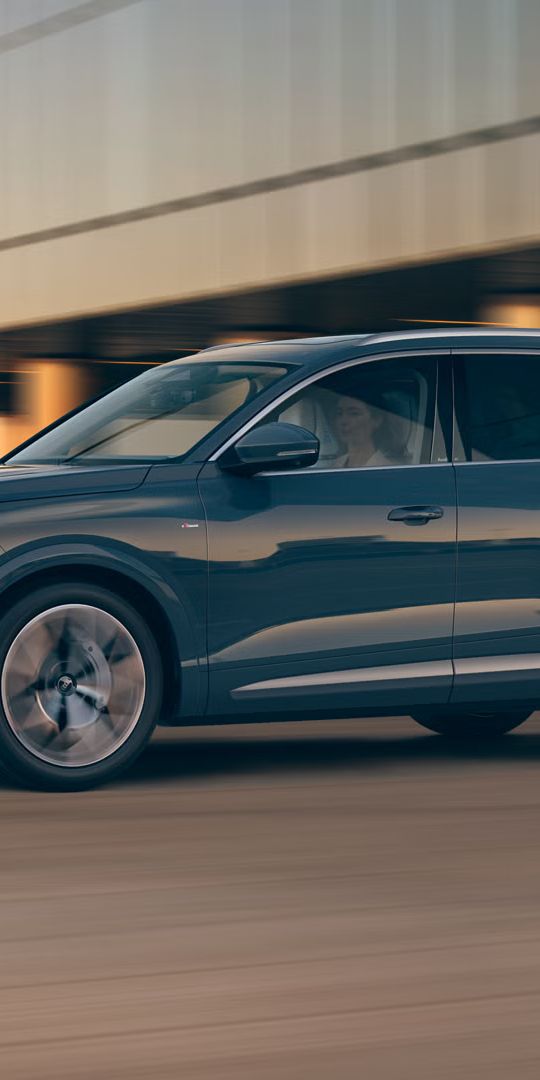 Modré SUV Audi Q7 ide po ceste pri západe slnka, v pozadí je moderná budova stojaca na betónových pilieroch.