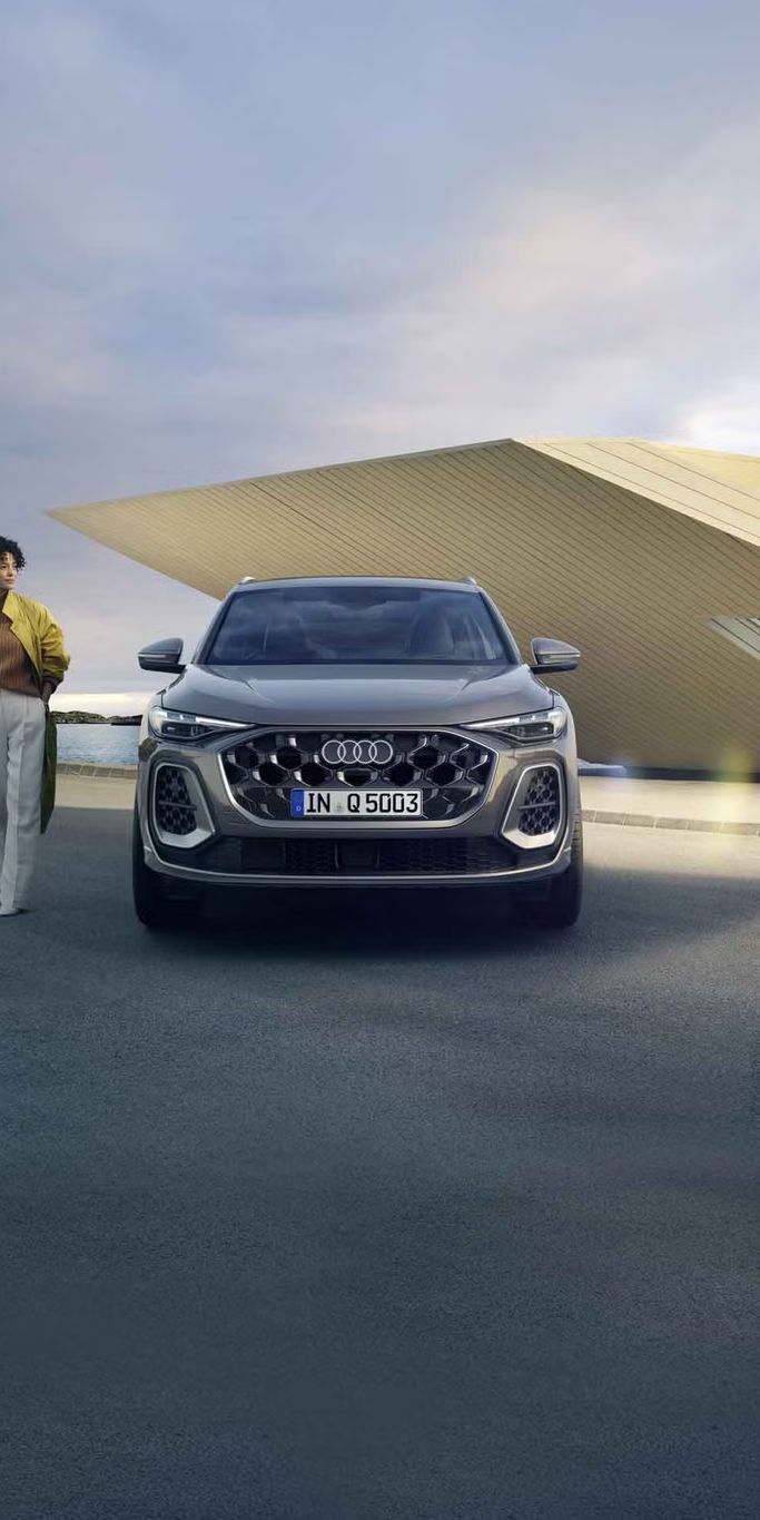 Der Q5 SUV im Frontprofil auf einem Parkplatz direkt an der Küste, dahinter ein kleines, futuristisches Haus. Neben dem Q5 SUV steht eine junge, modisch gekleidete Frau.	