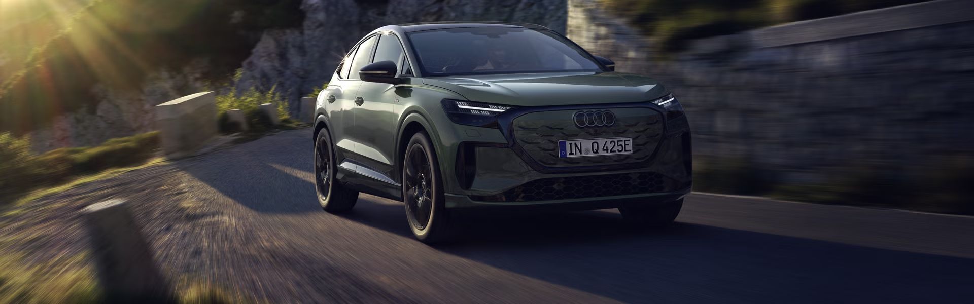 Q4 Sportback e-tron počas jazdy po horskej ceste. Slnečné lúče zľava hore osvetľujú obraz, vozidlo vrhá tieň dopredu.