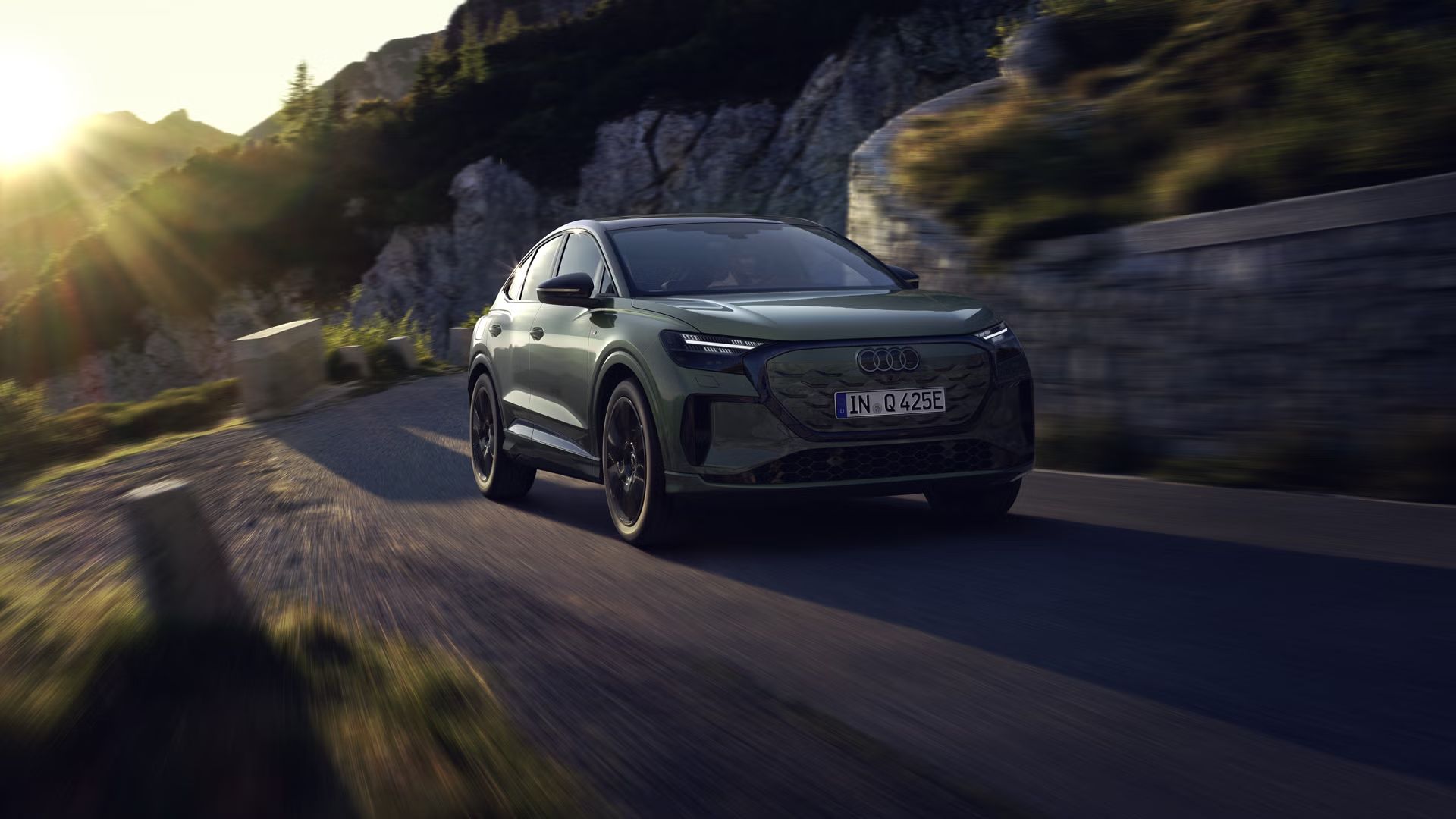 Q4 Sportback e-tron počas jazdy po horskej ceste. Slnečné lúče zľava hore osvetľujú obraz, vozidlo vrhá tieň dopredu.