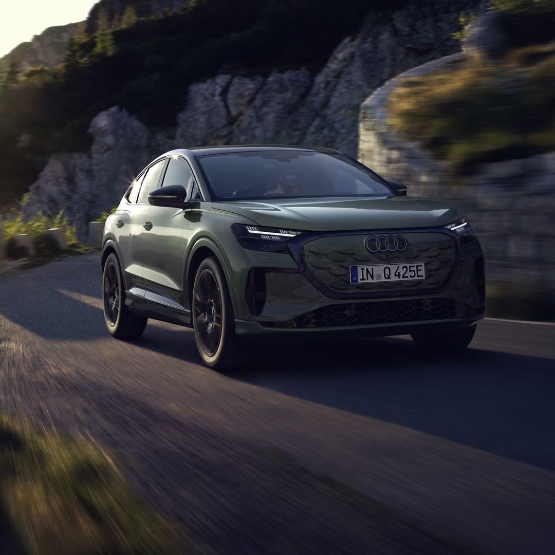 Q4 Sportback e-tron počas jazdy po horskej ceste. Slnečné lúče zľava hore osvetľujú obraz, vozidlo vrhá tieň dopredu.