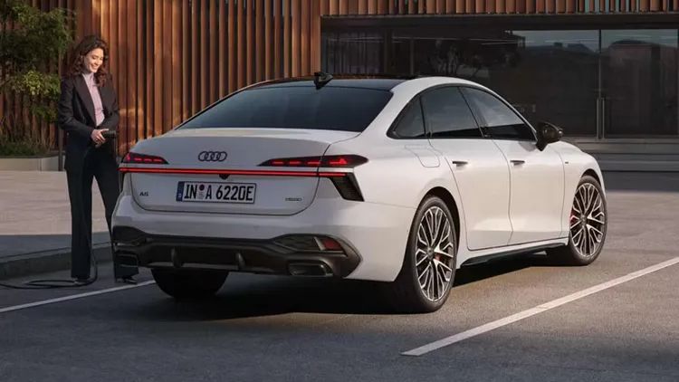 Biele Audi A6 Limuzína e-hybrid zaparkované na parkovisku pred modernou budovou so sklenenou fasádou s drevenými lamelami pred sklenenými plochami. Vedľa auta stojí žena v kostýme.