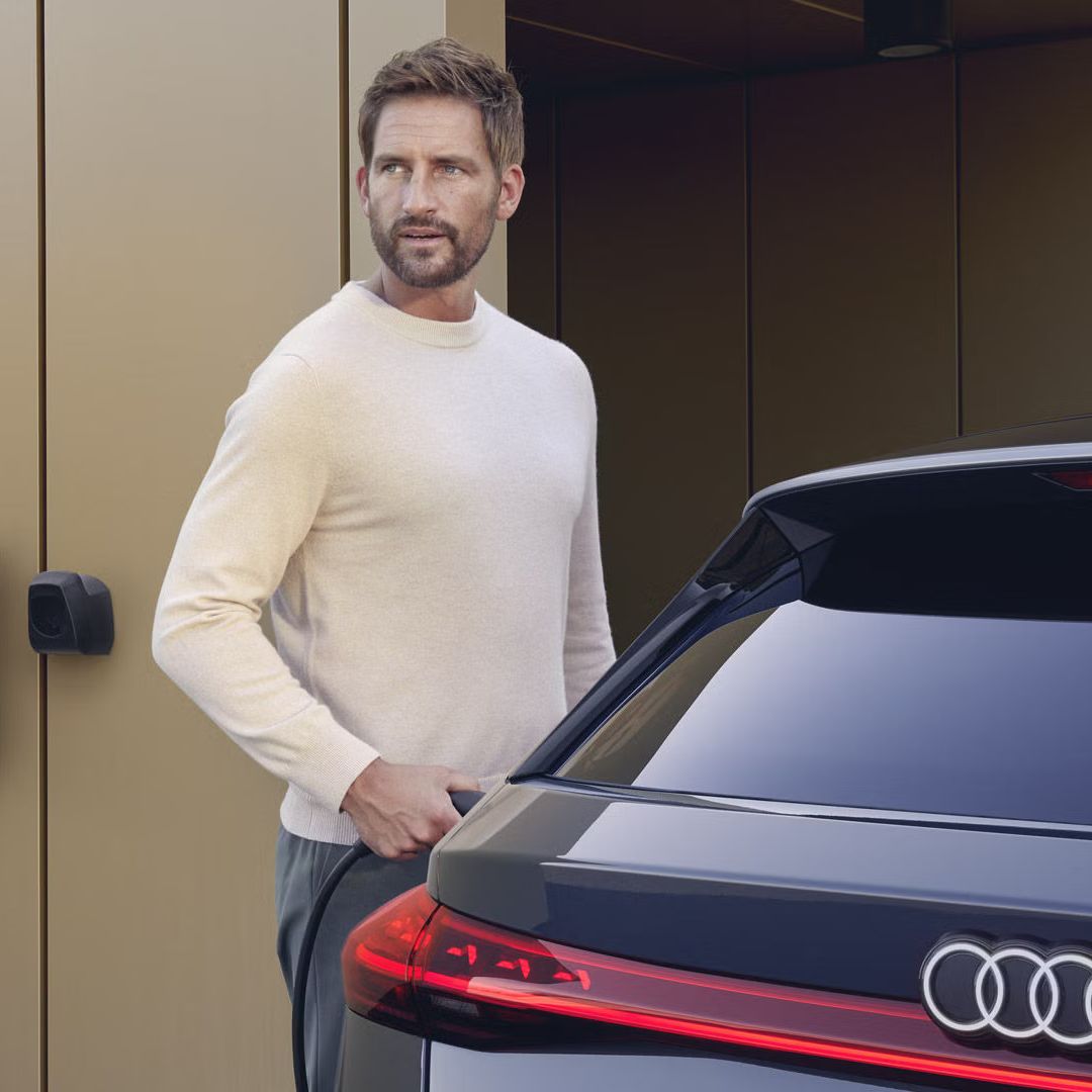 S6 Avant e-tron stojí pred moderným domom. Na fasáde domu je namontovaná nabíjací stanica, ktorú k vozidlu pripája muž stredného veku.