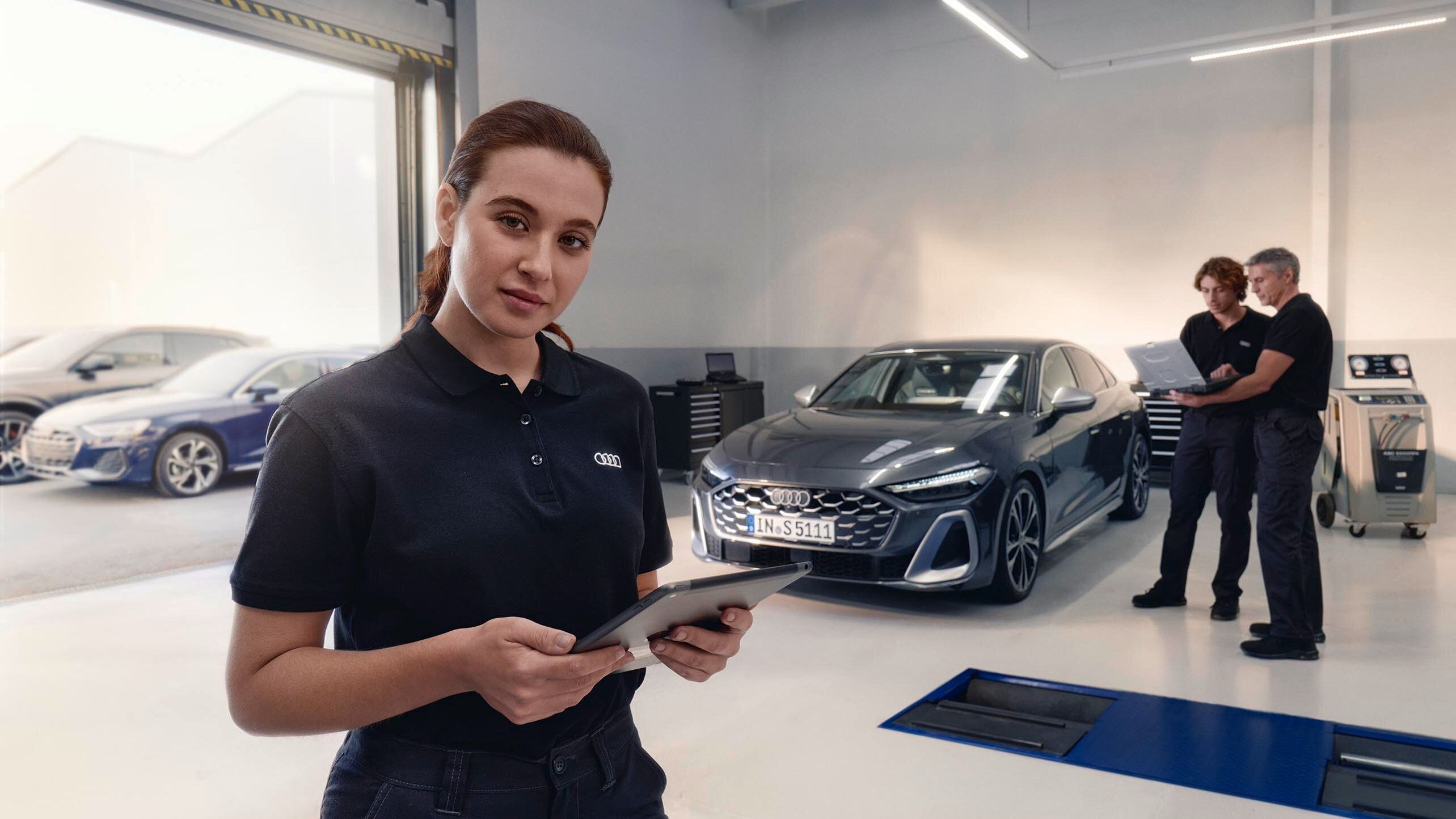 Servisná pracovníčka stojí v servise automobilov a drží tablet. V pozadí sú dvaja muži, ktorí sa rozprávajú pred zaparkovaným autom. Miestnosť je moderná a dobre osvetlená.