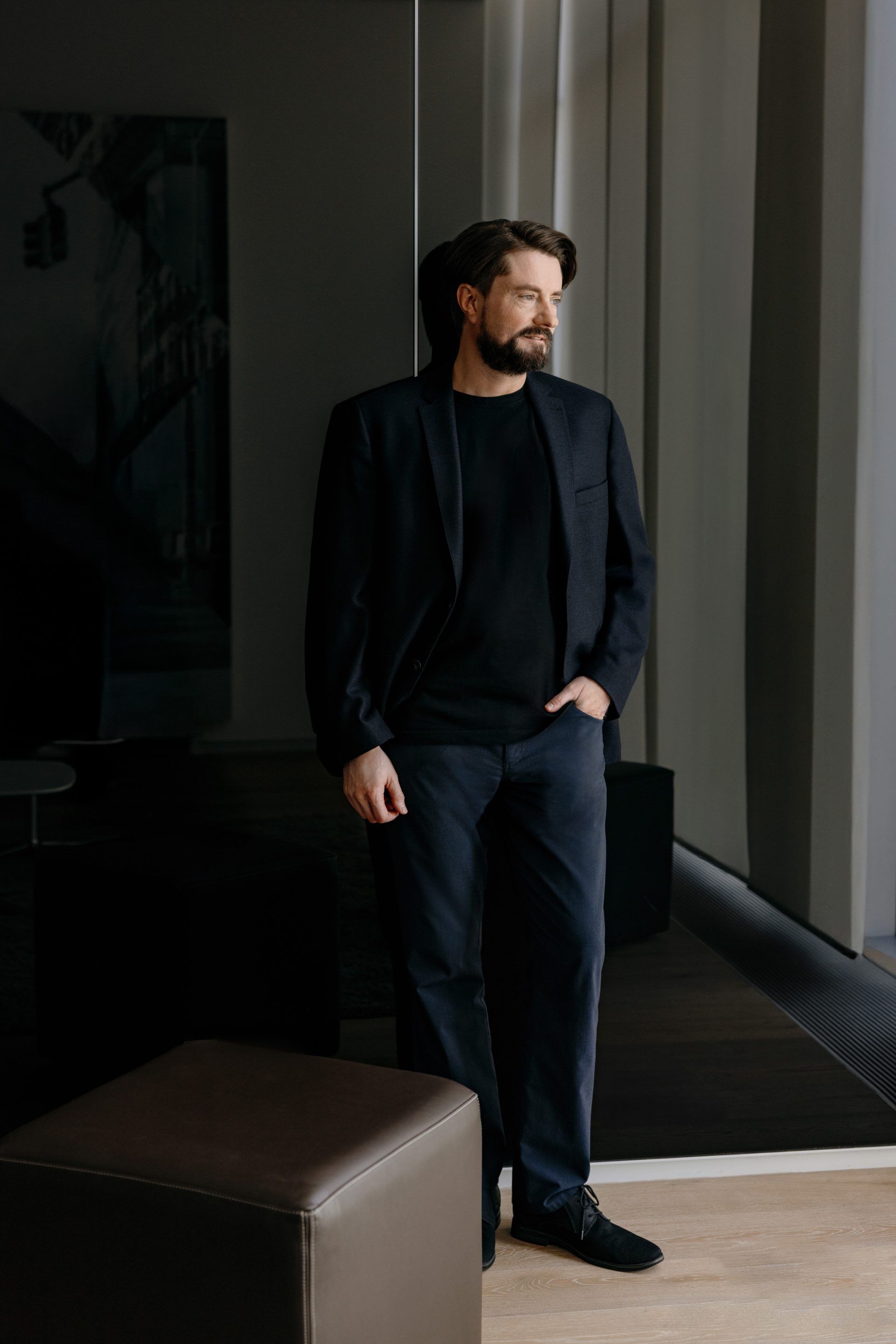 Muž oblečený v tmavom saku a nohaviciach stojí vedľa veľkého okna. Pozadie je tmavé, miestami osvetlené prirodzeným svetlom. Na zemi je viditeľná časť hnedého dizajnového nábytku.