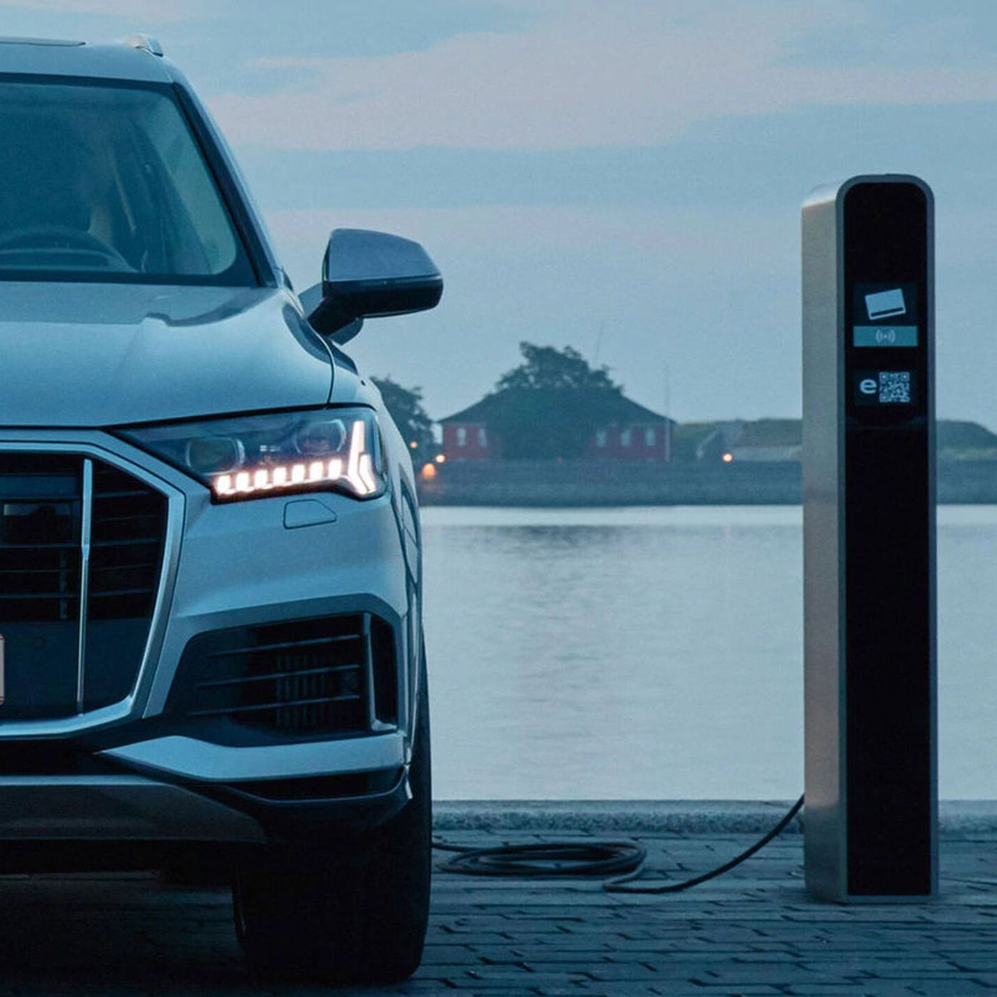 Elektrické SUV je zaparkované pri nabíjacej stanici na brehu vodnej nádrže. Na pozadí sú viditeľné domy a stromy. Auto sa nabíja pomocou kábla pripojeného k nabíjacej stanici..
