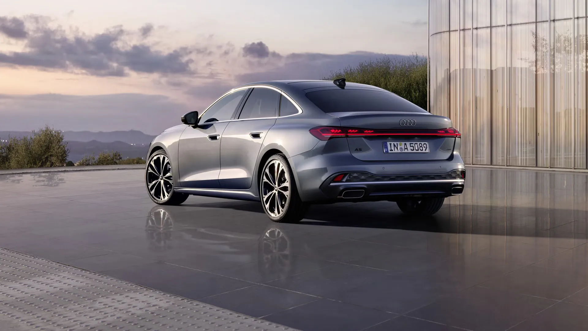 Audi A5 Limuzína zo šikmého pohľadu na zadné partie. Vozidlo sa odráža na hladkom povrchu. V pozadí sú viditeľné obrysy hôr za súmraku.