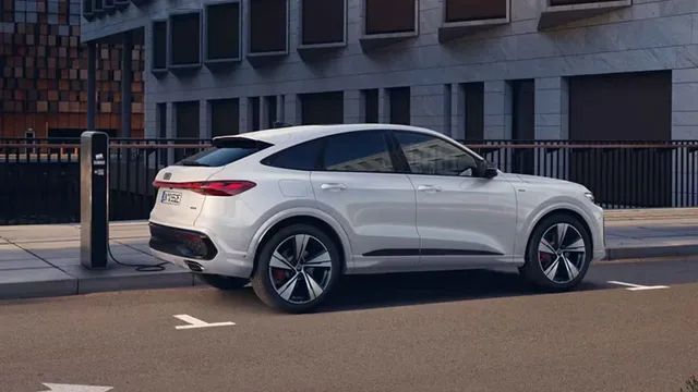 Moderné biele SUV zaparkované pri chodníku pred budovou s veľkými oknami. Auto má športový dizajn a elegantné línie. Vozidlo sa nabíja na nabíjacej stanici.