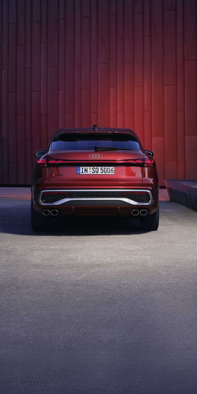 Heckprofil des roten SQ5 Sportback, welcher in einem Stadtpark vor einer Gebäudefassade steht und diese rot anleuchtet.