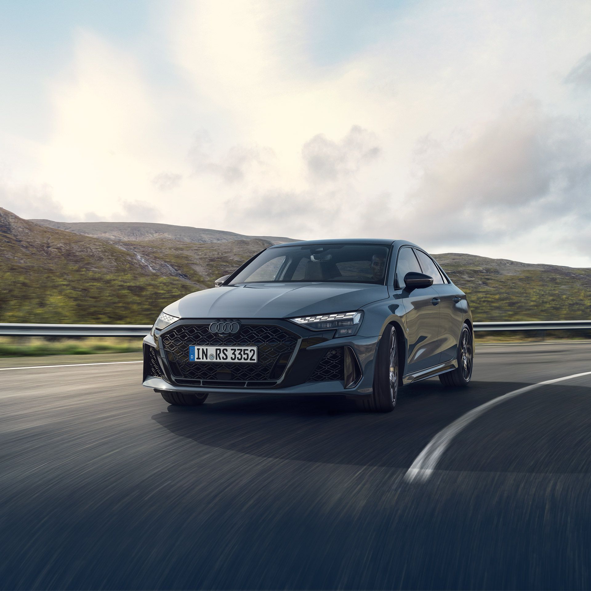 Športové Audi RS3 jazdí po kľukatej horskej ceste za slnečného počasia. Pozadie tvoria hory a jemne zatiahnutá obloha s niekoľkými oblakmi. Auto ide dynamickým štýlom, čo zdôrazňuje jeho výkon.