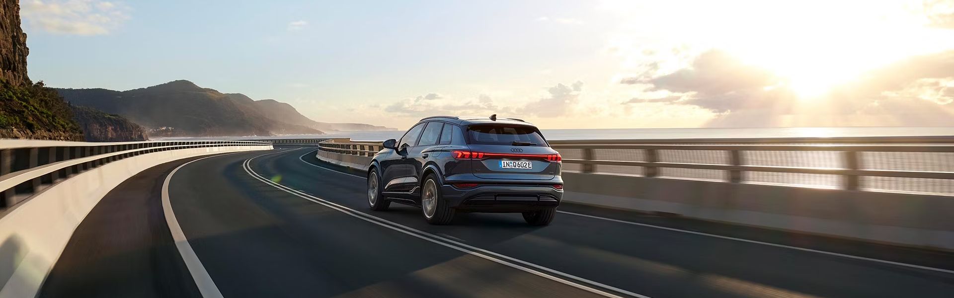 Pohľad na zadné partie Audi Q6 počas dynamickej jazdy po ceste popri pobreží.