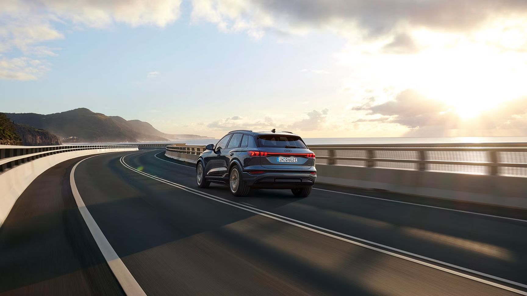 Pohľad na zadné partie Audi Q6 počas dynamickej jazdy po ceste popri pobreží.