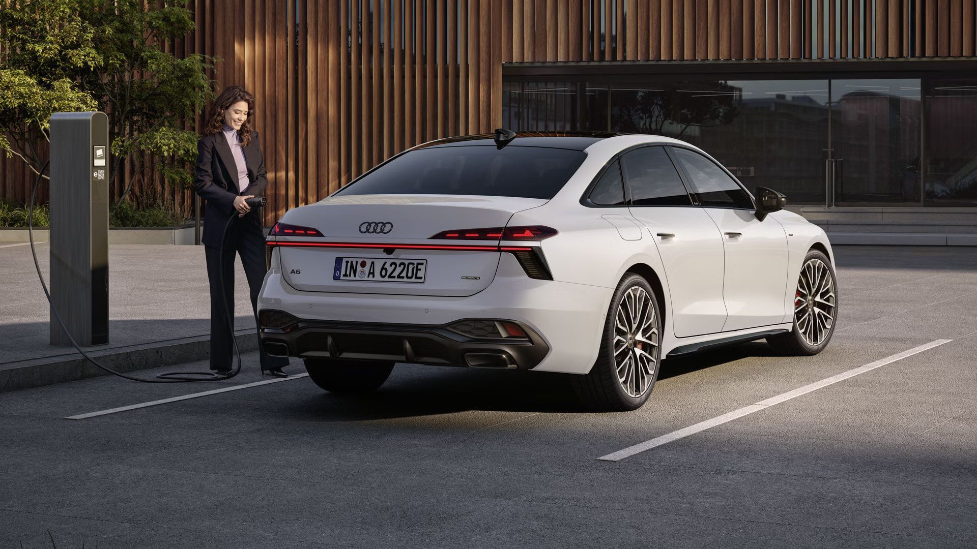 Biele Audi A6 e-hybrid  stojí na parkovisku pred budovou. Mladá žena pripája Audi A6 Limuzína e-hybrid k nabíjacej stanici.