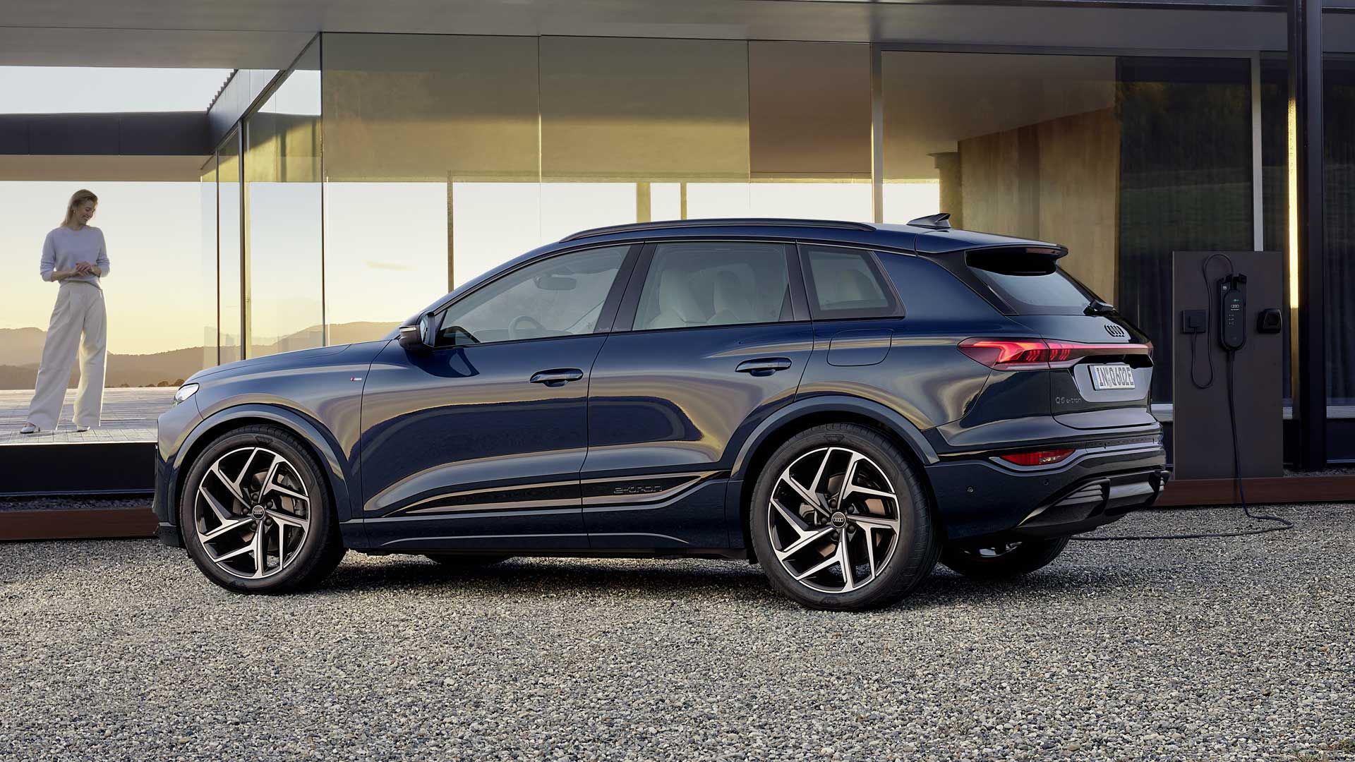 Audi Q6 SUV e-tronstojí na štrkovom povrchu pred modernou vilou so sklenenou fasádou. Pred domom stojí mladá žena vo svetlom oblečení a pozerá sa na vozidlo, ktoré je napojené na nabíjaciu domácu stanici a nabíja sa. 