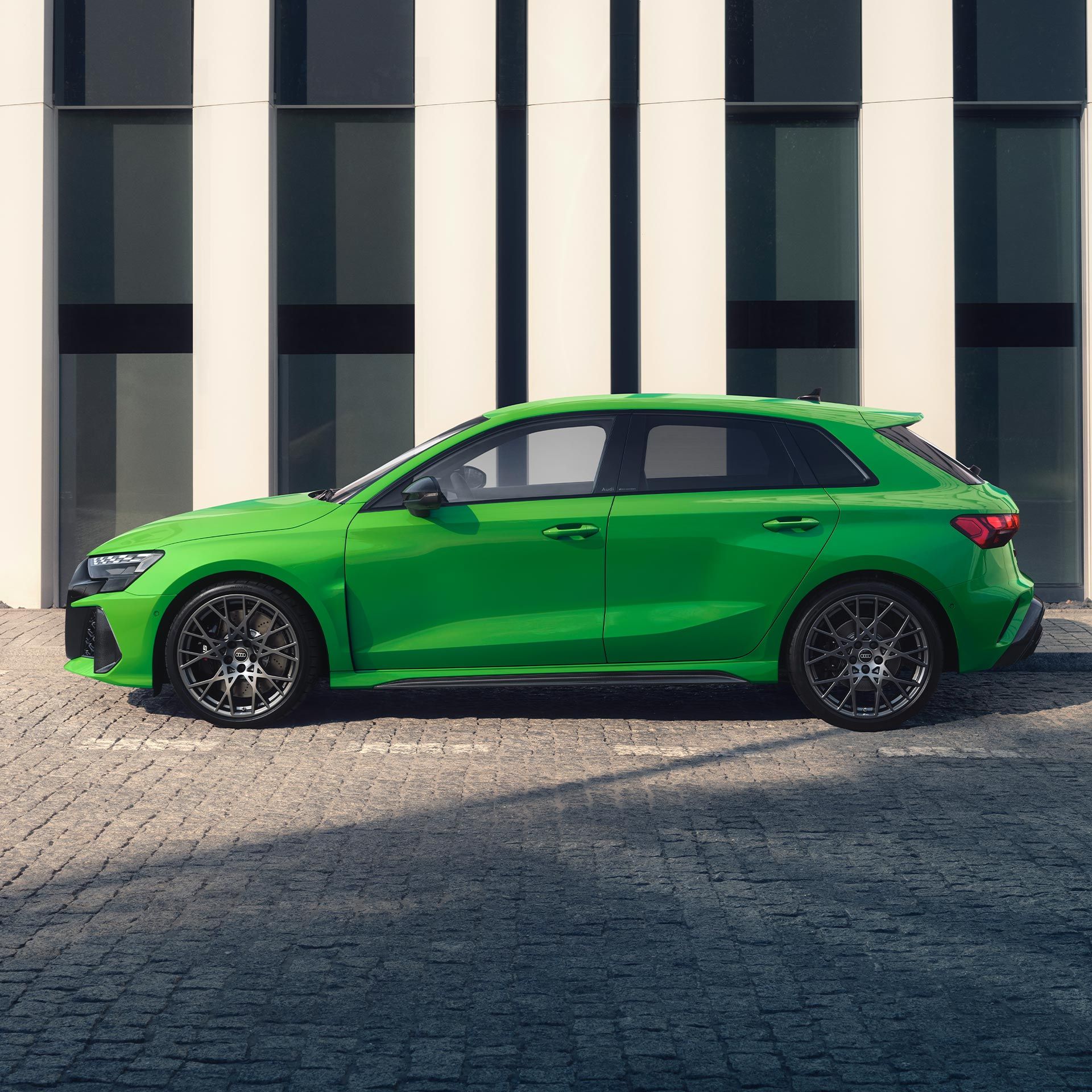 Zelené Audi RS3 Sportback zaparkované na kamennej dlažbe pred modernou budovou.