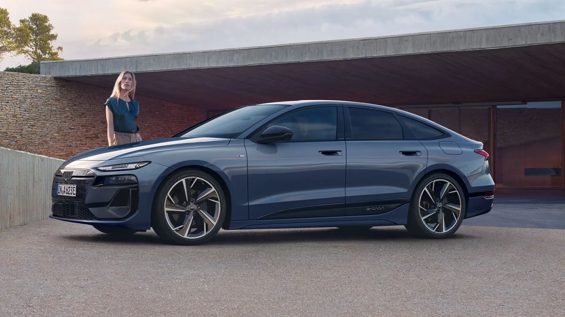 Audi A6 Sportback e-tron je zaparkovaný pred moderným domom. Vedľa vozidla stojí mladá žena s blond vlasmi.
