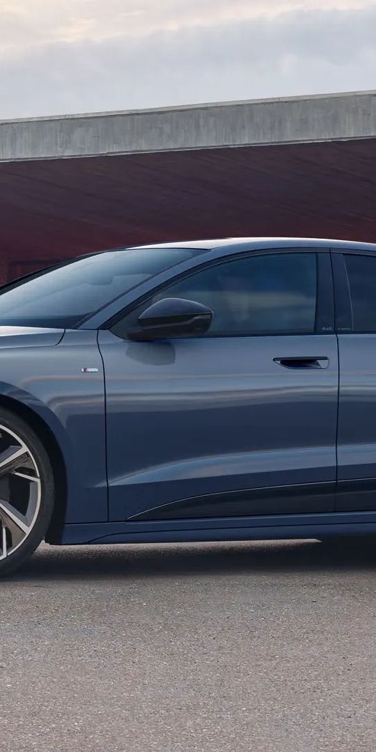 Audi A6 Sportback e-tron je zaparkovaný pred moderným domom. Vedľa vozidla stojí mladá žena s blond vlasmi.
