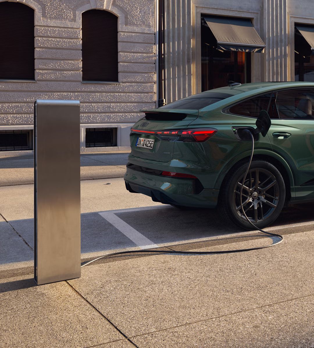 Pohľad na pravú zadnú časť vozidla Q4 Sportback e-tron, zaparkovaného pri nabíjacej stanici. V pozadí je moderná budova. Kábel nabíjacej stanice je pripojený k vozidlu.