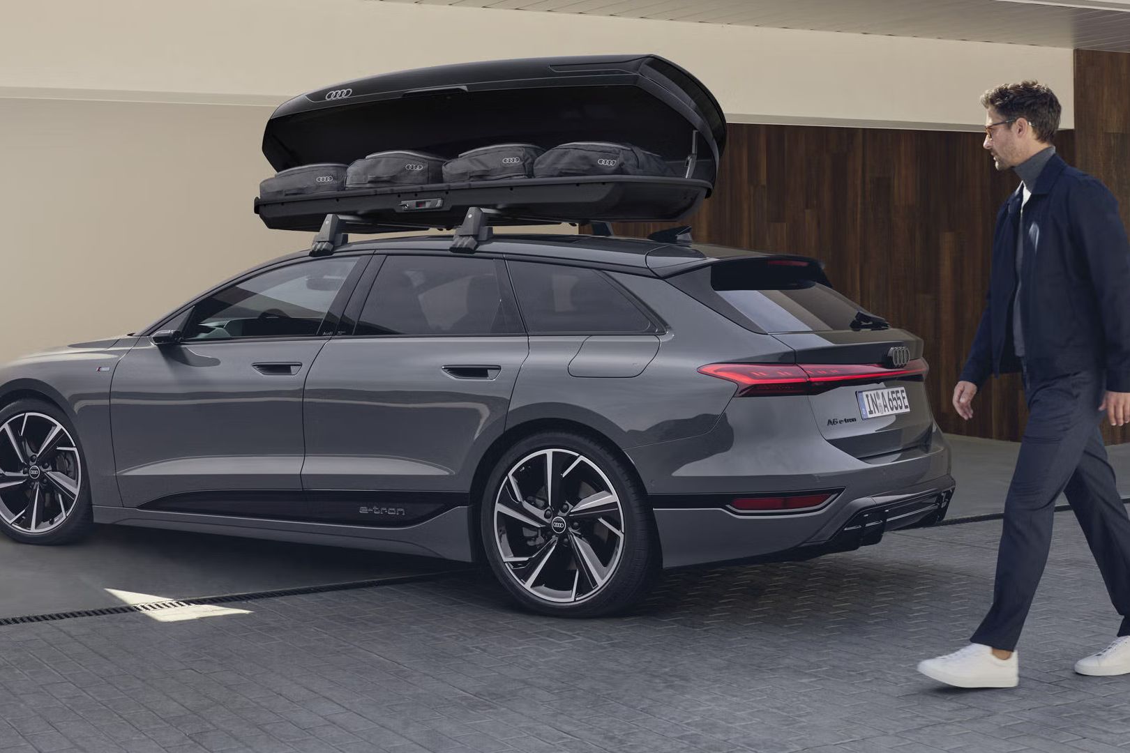 A6 Avant e-tron stojí šikmo pred moderným domom, k vozidlu sa blíži muž. Na streche je namontovaný strešný box s otvoreným vekom. V strešnom boxe sú viditeľné tašky. 