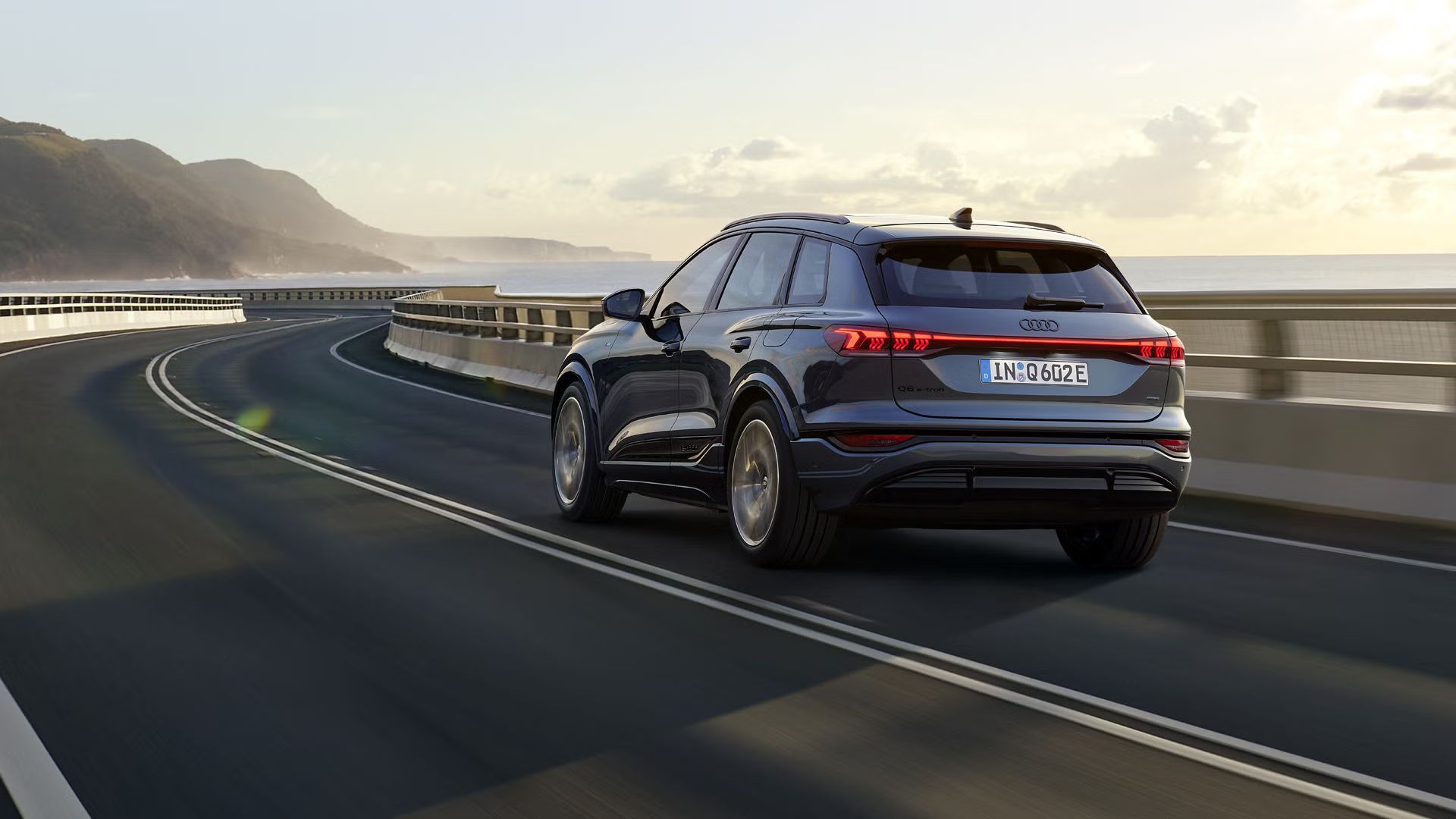 Pohľad na zadné partie Q6 SUV e-tron, ktoré jazdí po kľukatej ceste pri mori. V pozadí sú vidieť hory.