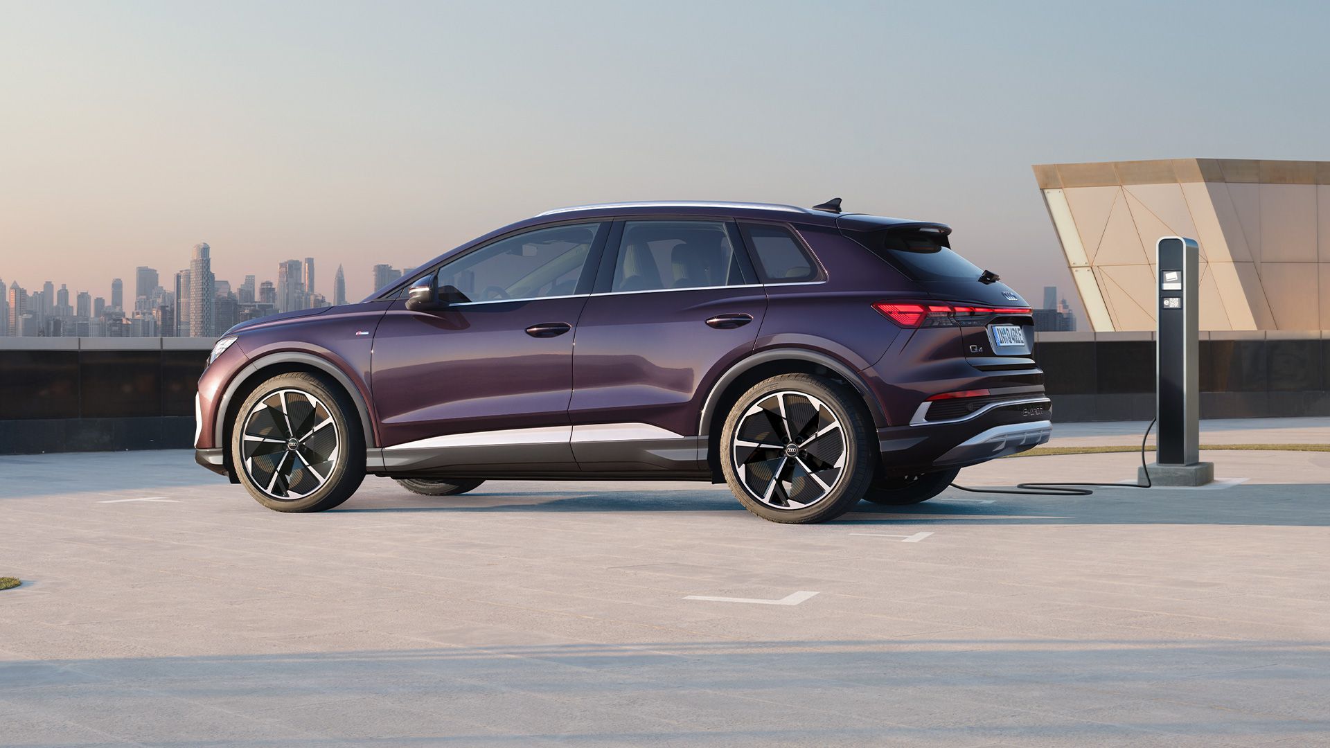 Audi Q4 e-tron zaparkované na parkovacom mieste pri verejnej nabíjacej stanici. Vozidlo sa nabíja. V pozadí je moderná budova a výhľad na centrum mesta s mrakodrapmi.