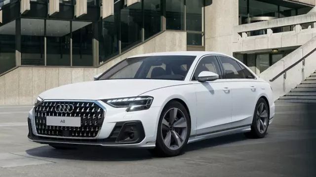Luxusné biele Audi A8 TFSI e stoji pred modernou budovou. Auto má športový dizajn s charakteristickou jednodielnou maskou chladiča. V pozadí moderná, mestská betónová budova.