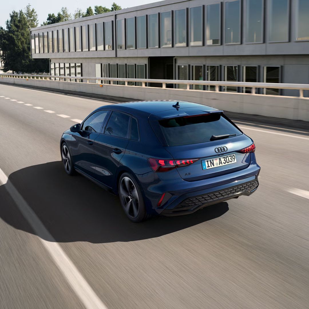 Audi A3 Sportback zo zadného pohľadu na moste, pričom dopadajúce slnko vrhá veľký tieň na ľavú stranu vozidla. V pozadí kancelárska budova.